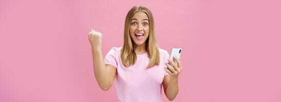 Girl beat own record in smartphone game raising clenched fist in cheer and triumph holding cellphone, smiling excited and happy at camera celebrating victory with joyful gesture over pink background photo