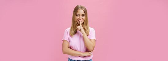 Shh keep it secret. Portrait of charismatic cheerful chubby girl with tanned skin and fair hair showing shush gesture with index finger over mouth smiling hiding surprise posing over pink background photo