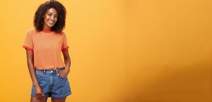 Ready to travel world. Energetic confident and attractive dark-skinned woman with curly hair holding hand in pocket of denim shorts smiling joyfully posing over orange background carefree and friendly photo