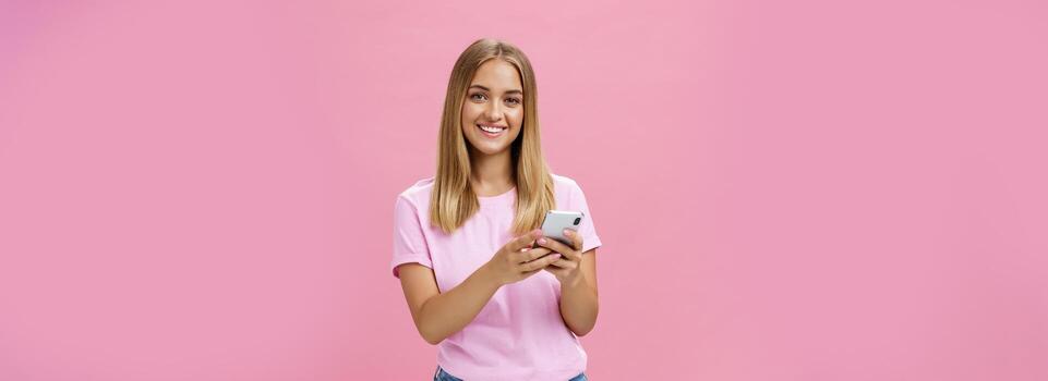 Woman calling taxy via smartphone asking friend address smiling cheerfully at camera holding phone with both hands over chest getting in touch with clients via messages standing over pink wall photo