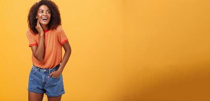 Stylish feminine and fashionable african american female model with afro hairstyle touching neck gently looking right with amused carefree expression holding hand in pocket over orange background photo