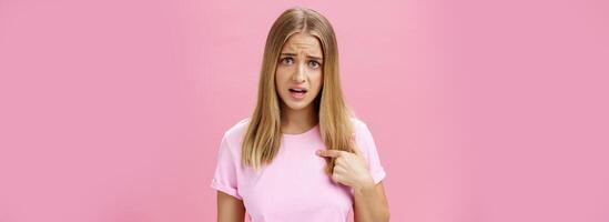 Insulted girl pointing at herself with displeased pissed and questioned expression asking question being shocked she picked or accused in something terrible and disrespectful posing against pink wall photo