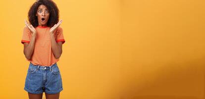 Oh no what happened. Worried african american sister with curly hair in trendy striped t-shirt and denim shorts gasping dropping jaw, looking concerned expressing empathy and shock over orange wall photo