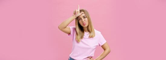 Girl mocking under boyfriend showing loser sign on forehead sticking out tongue gazing arrogant and indifferent at camera being moody holding hand on waist posing against pink background photo