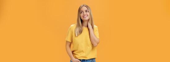 Pretty and tender cute european female independent freelancer in yellow t-shirt sighing touching neck and gazing dreamy at upper right corner with pleasant smile, posing over orange background photo
