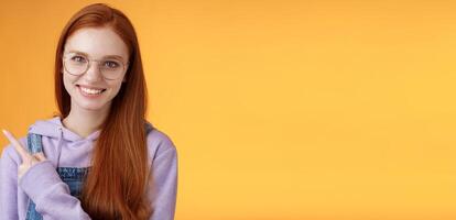 Friendly good-looking modern redhead young girl pointing left index finger showing awesome place suggesting go hang out smiling joyfully casually talking discuss new product, orange background photo