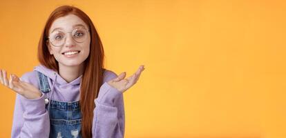 Well no big deal. Attractive silly redhead hipster girl wearing glasses smiling awkward unaware spread hands sideways shrugging confused grinning questioned uncertain what tell, orange background photo