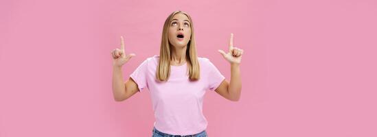 retrato de divertido y impresionado curioso atractivo mujer en camiseta goteante mandíbula mirando y señalando arriba asombrado y intrigado acecho interesante objeto en cielo posando en contra rosado antecedentes foto