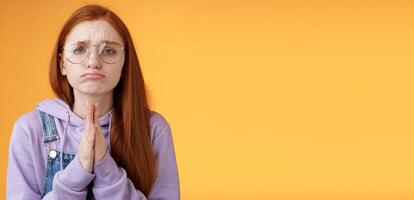 Please beg you. Clingy upset sad caucasian redhead girlfriend wearing glasses frowning grimacing pouting pleading help asking favour need urgently borrow something, orange background photo