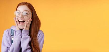 Excited happy smiling broadly redhead girlfriend drop jaw grinning thrilled look left astonished touch cheeks reacting awesome stunning news screaming happiness, standing orange background photo