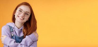 Tender romantic european redhead girlfriend tilting head cute lovely smile white teeth tilting head hugging herself wearing best friends hoodie adore new outfit grinning joyfully, orange background photo