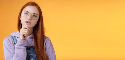 Thoughtful young creative smart redhead girl thinking figure out important thing standing upper left corner squinting thinking get clue touch chin thinking, pondering choice, orange background photo