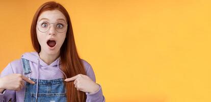 Shocked emotive surprised redhead european woman wearing glasses hoodie drop jaw gasping stunned pointing herself full disbelief cannot believe win being picked look impressed orange background photo