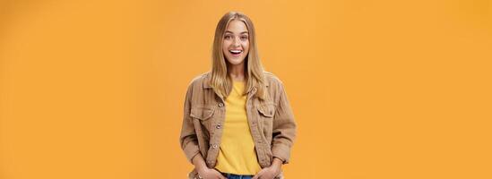 Charismatic tanned woman in corduroy jacket and yellow t-shirt ready for chilly autumn walk with friends smiling joyfully gazing entertained at camera holding hand in pockets casually over orange wall photo