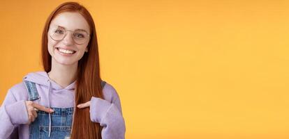 Happy lively friendly redhead european girl pointing herself suggesting help boasting telling own accomplishments smiling white teeth delighted volunteering, being picked, orange background photo