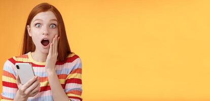 Concerned shocked emotive redhead girl find out stunning news look camera drop jaw gasping impressed touch cheek hold smartphone finish reading impressive disturbing story, orange background photo