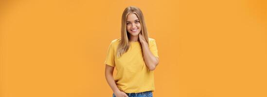 retrato de tímido y tímido femenino niña con bronceado y Derecho hecho pelo frotamiento cuello y sonriente sensualmente con contento despreocupado expresión participación mano en bolsillo posando en contra naranja antecedentes foto