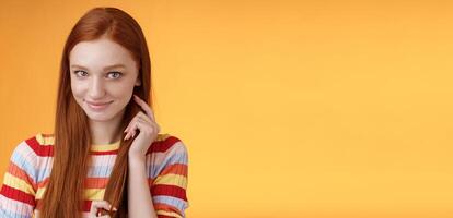 Romantic flirty shy attractive redhead girl 20s touching hair strand smiling silly modest glancing camera coquettish making lovely glances wanna seduce guy expressing sympathy, orange background photo