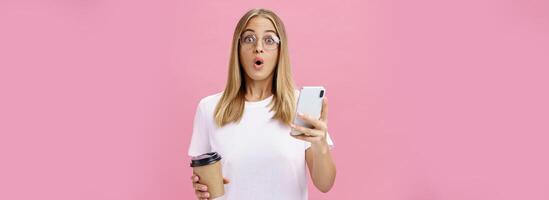 Girl receiving awesome message on smartphone while drinking coffee from cup in cafe sharing news with friend folding lips in wow sound staring surprised at camera holding cellphone over pink wall photo