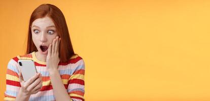 Omg what heck. Portrait shocked concerned young redhead sensitive impressed redhead woman stare smartphone display touch cheek drop jaw stunned surprised standing orange background hold phone photo
