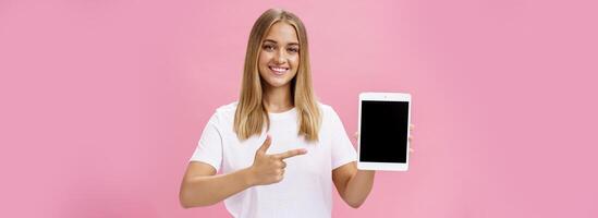 niña recomienda utilizar solamente esta digital tableta. carismático joven hembra con justa pelo bronceado piel y No maquillaje en blanco camiseta señalando a artilugio pantalla y sonriente simpático a cámara terminado rosado pared foto