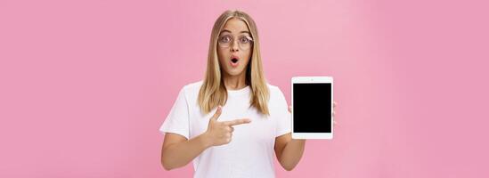 Shocked intelligent cute female in glasses and white t-shirt folding lips from amazement in wow sound gasping popping eyes amused holding digital tablet pointing at gadget screen over pink wall photo