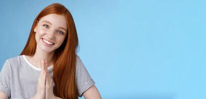 Cheerful sincere kind redhead girl do coquettish eyes tilting head look nice asking favour say please smiling tender press palms together supplicating pleading begging help, standing blue background photo