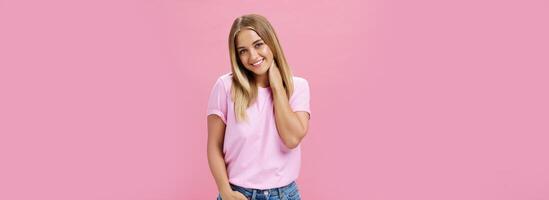 Portrait of shy and timid attractive european woman with fair hair in casual outfit touching neck silly tilting head and smiling cute at camera standing over pink background photo