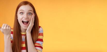 Cheerful excited impressed talkative girl pointing astonished thumb left drop jaw smiling broadly touch cheek thrilled, interesting event discuss awesome wonderful performance, orange background photo