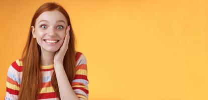 Cheerful glad excited attractive redhead girl blushing surprised feeling happy touch cheek pleased receive good news standing joyful thrilled get awesome chance, posing orange background photo