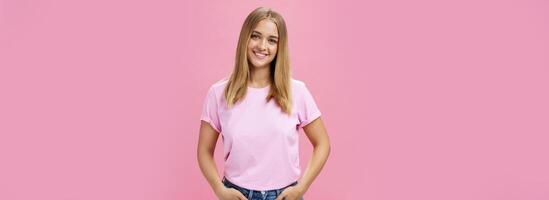 encantador agradable mujer con gordito cara y justa pelo en casual camiseta sonriente en general participación manos en bolsillos siendo bonito y simpático mirando despreocupado y calma a cámara terminado rosado antecedentes foto