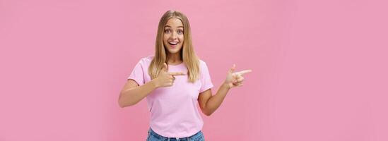 Portrait of charismatic and enthusiastic good-looking female with blonde hair and tanned skin in casual t-shirt pointing left smiling amused and delighted standing impressed over pink background photo