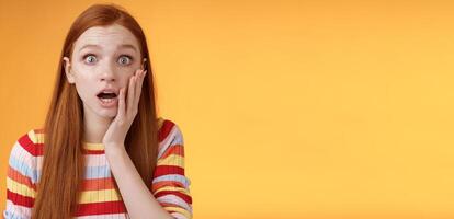 Concerned worried ginger girl blue eyes drop jaw gasping touch cheek confused looking nervously anxious, show empathy hearing terrible upsetting story standing orange background photo