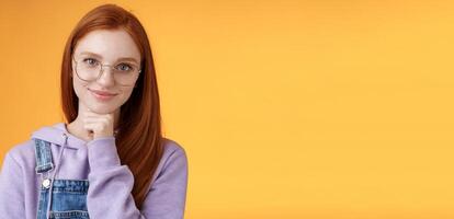 Confident happy relaxed european redhead skillful young female entrepreneur glasses establish startup smiling delighted look pleased result touch chin thoughtful smart, orange background photo