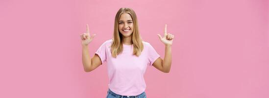 Indoor shot of pleasant attractive friendly-looking girl with tanned skin in casual t-shirt and jeans raising hands pointing up and smiling broadly at camera with satisfied look over pink background photo