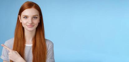 Attractive modest good-looking redhead girlfriend acting mature friendly smiling pointing left index finger giving direction show way bathroom standing blue background joyful kind grin photo