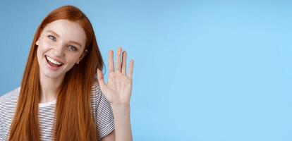 Attractive confident redhead sassy girl pure clean skin blue eyes tilting head cheerfully waving hand hello hi gesture greeting you look camera friendly welcoming friend, standing studio background photo