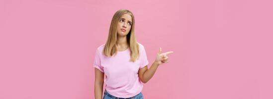 Envy upset cute young european woman with tanned skin and fair hait tilting head lifting eyebrows in sad silly look pointing, gazing left with regret and disappointment posing against pink background photo