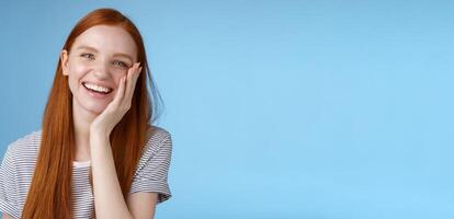 Charismatic talkative friendly-looking happy laughing redhead girl having fun discuss previous summer holidays make jokes chuckling touching face amused standing cheerful blue background photo