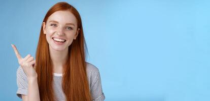 Happy charismatic redhead laughing young girl having fun looking carefree talking discussing new product sale pointing up index finger showing copy space advertisement, standing blue background photo