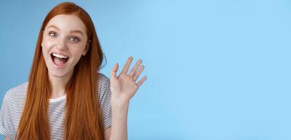 Hello wanna be friends. Enthusiastic cute redhead female newbie getting know coworkers smiling happy waving raised hand hi greeting gesture welcoming, say bye standing blue background photo
