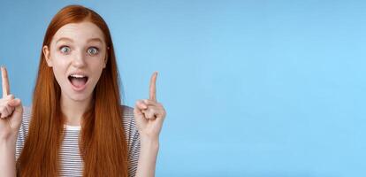 Surprised happy enthusiastic young redhead woman reacting impressed upper hanging promo pointing up index fingers drop jaw amused look thrilled camera telling about awesome offer, blue background photo