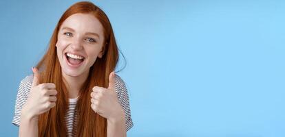 Enthusiastic amused outgoing redhead beautiful girl say yeah supportive like awesome idea show thumbs-up approval recommend gesture agree good choice, standing blue background photo