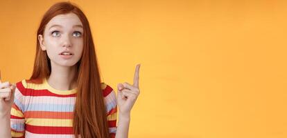 Stunned thrilled young redhead woman peer focused pointing up index fingers upwards look concentrated excited hold breath amused performance standing orange background intrigued and curious photo