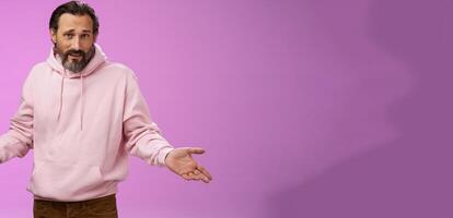 Bothered confused mature adult bearded man grey hair in pink hoodie arguing look pissed offended shrugging spread hands sideways dismay look clueless camera asking why, purple background photo