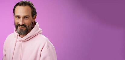 Charming alluring handsome bearded stylish adult male model earring pink hoodie smiling delighted express confidence positivity feel lucky amused, standing purple background talking casually photo