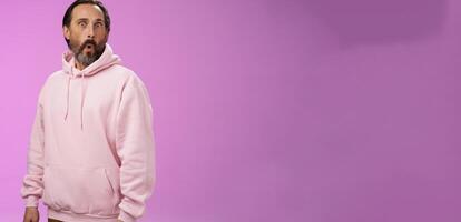 Impressed speechless shocked adult bearded man grey hair in cool pink hoodie folding lips wow popping eyes camera surprised astonished intrigued, standing curious amazed purple background photo