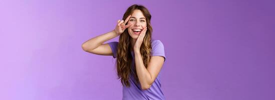 Adorable lovely funny cute woman curly brown hairstyle tilt head touch cheek blishing silly flirty smiling camera show peace victory sign express positive joyful attitude stand purple background photo