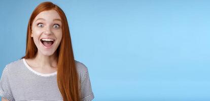 Surprised pleased happy impressed redhead european girl 20s reacting amused wide eyes look admiration joy receive incredible offer standing excited express thrill upbeat feelings, blue background photo