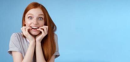 Excited amazed attractive redhead girl blue eyes look fascinated affection smiling desire bite fingernails eager feel astonished cannot wait bite tasty food, standing blue background thrilled photo